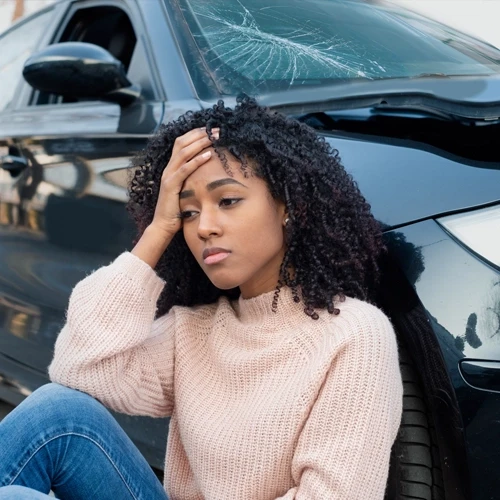 A young woman holding her head in pain and leaning against her damaged car after an accident
