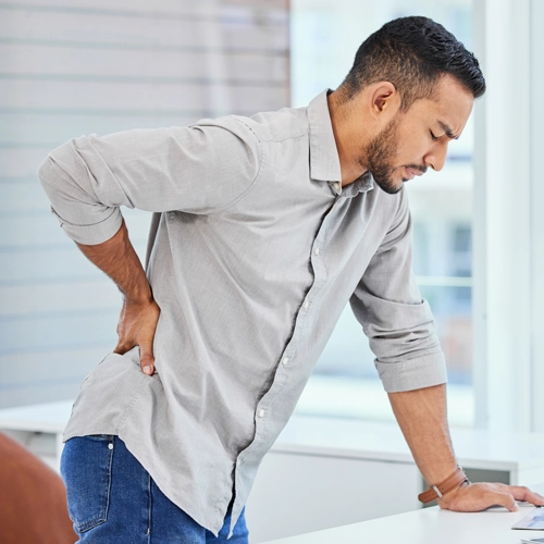 A man standing up from his desk and holding onto his lower back in pain